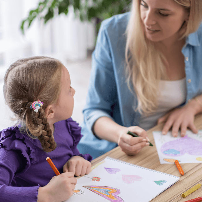 Adult and child sit at a table drawing colorful pictures with crayons, including a rainbow and hearts, while talking together.